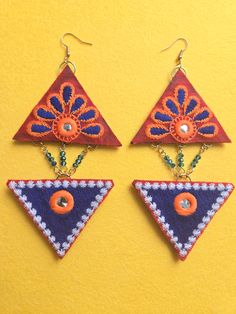 Beaded Blue Petal Embroidered Triangle Earrings Two embroidered fabric stacked triangle shapes arranged in a diamond shape dangle earring. Blue glass beaded chain between triangle shapes. Top triangle has a red background with blue and orange petals surrounding a small circular mirrored center. The bottom triangle is mostly blue with white edges and a small circular mirrored center. Earrings are 5 inches long and 2.5 inches wide with gold 18k gold plated ear wires.