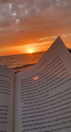 an open book sitting on top of a sandy beach next to the ocean at sunset