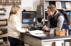 a man and woman sitting at a desk in front of a computer monitor talking to each other