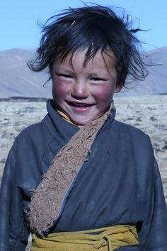 a young boy with black hair and blue eyes smiles at the camera while standing in an open field