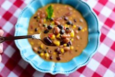 a person is holding a spoon over a bowl of beans and corn soup on a checkered tablecloth