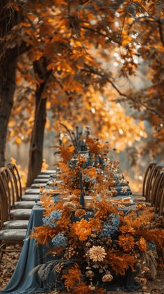 a long table with blue cloths and orange flowers on it in the middle of some trees