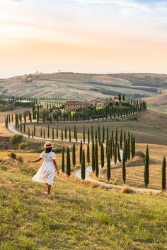 a woman in a white dress and hat is walking on a hill with cypress trees