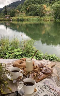 two cups of coffee are sitting on a picnic table near the water and some bread