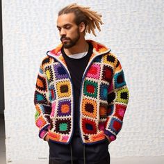 a man with dreadlocks standing in front of a wall wearing a multicolored jacket