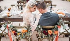 a bride and groom sitting at a table with flowers on the back of their chairs