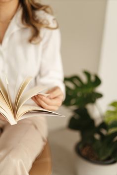 a woman reading a book while sitting on a chair next to a potted plant