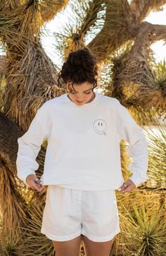 a woman standing in front of a palm tree with her hands on her hips wearing a white sweatshirt and shorts