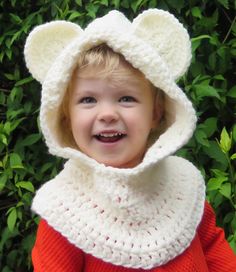 a young child wearing a crocheted bear hat and scarf in front of some bushes