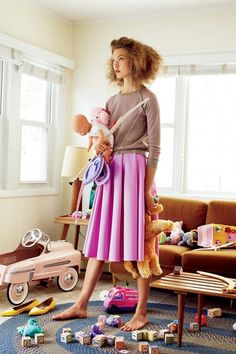 a woman in a pink skirt holding a baby doll next to a living room filled with toys