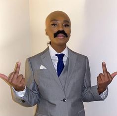 a man wearing a suit and tie with two fingers up in the air while standing next to a white wall