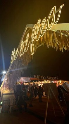 people are gathered under the neon sign at an outdoor event that is lit up with lights and streamers