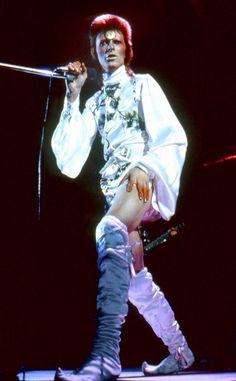 the man is dressed in all white and holding a microphone while standing on stage with his legs crossed