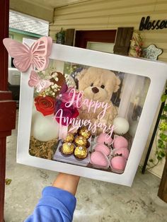 a person holding up a happy anniversary sign with chocolates and teddy bear in it