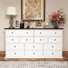 a white dresser with drawers and vases on top next to a lamp, rug and painting