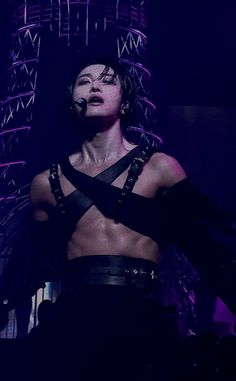 a woman with no shirt on standing in front of a purple lit background and wearing harnesses