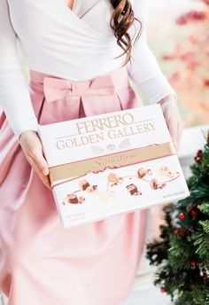 a woman is holding a box of golden gallerys in front of a christmas tree