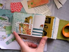 a person holding an open book on top of a table with papers and scissors next to it