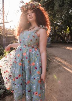 a woman wearing a dress with flowers on her head standing in front of some bushes