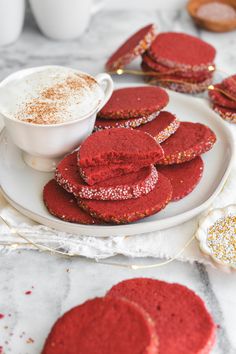 red velvet cookies on a white plate next to a cup of hot cocoa and sugar