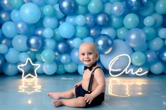 a baby sitting on the floor in front of blue balloons with one word written above it