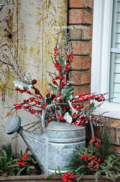 a metal watering can with red berries and greenery in front of a brick building