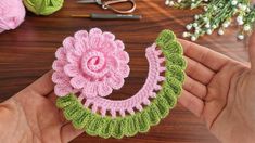 a hand holding a crocheted flower on top of a wooden table next to yarn and scissors