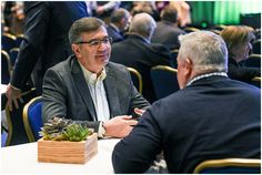 two men sitting at a table talking to each other in a room full of people