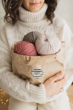 a woman holding a paper bag filled with balls of yarn and wearing a white sweater