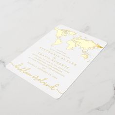 a white and gold wedding card with a world map in the background on a marble surface