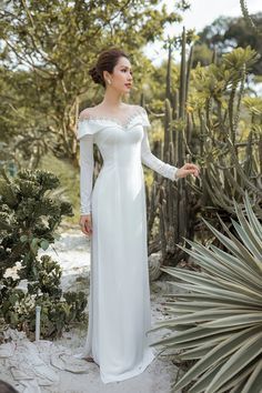 a woman in a white dress standing next to some cactus bushes and trees with her hand on her hip