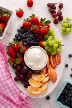 a white plate topped with fruit and dip surrounded by grapes, apples, oranges