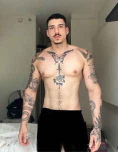 a man with tattoos on his chest standing in front of a bed and looking at the camera