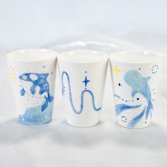 three ceramic cups with designs on them sitting on a white tablecloth, one is blue and the other has gold stars