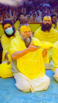 several men dressed in yellow sit on the ground and hold plates with their faces painted