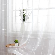 a vase with flowers on a table in front of a sheer curtained room window