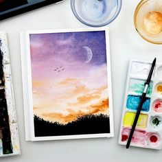 watercolors and paint supplies on a table with an open notebook, glass cup, and painting utensils