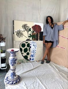 a woman standing next to some paintings on a white sheeted surface with flowers in vases