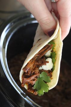 a hand holding a tortilla with meat and cilantro on it in a bowl