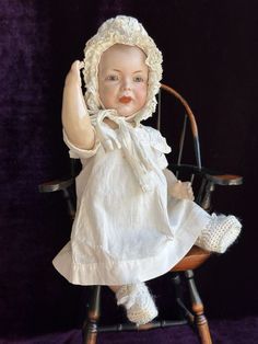 a doll sitting on top of a wooden chair with a white dress and bonnet over it's head