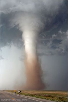 a large tornado cloud is seen in the sky over an open road with people walking on it