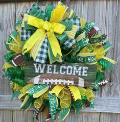 a football wreath with green and yellow ribbons hanging on a wooden fence that says welcome