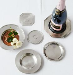 a bottle of champagne and three silver plates with flowers next to it on a white surface