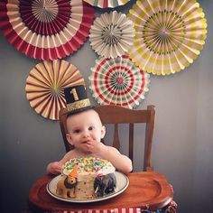 a baby sitting in a chair with a cake on it's lap and paper fans behind him