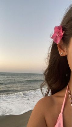 a woman standing on top of a beach next to the ocean with a pink flower in her hair