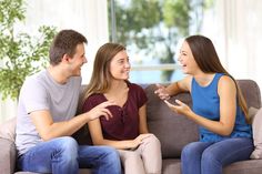 three people sitting on a couch laughing and talking to each other while one person points at something