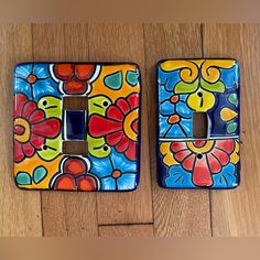 two colorful light switch plates on a wooden floor with flowers painted on the front and back