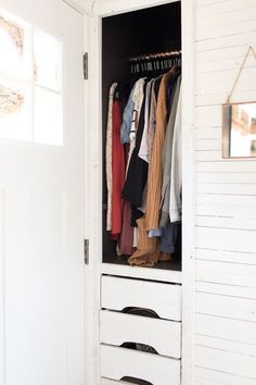 an open white closet with clothes hanging on the door and shoes in the bottom drawer