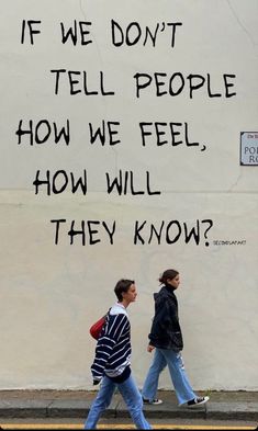 two people walking down the street in front of a wall with words written on it