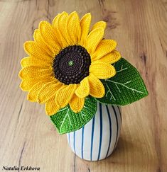 a crocheted sunflower in a white vase on a wooden table with green leaves